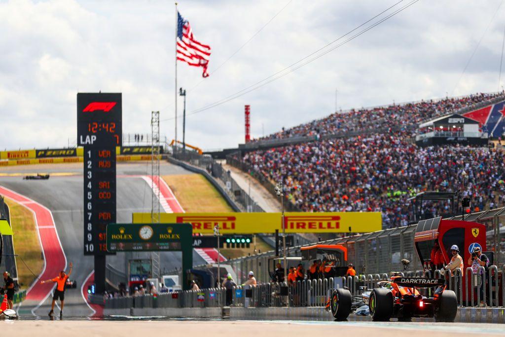 A rear view of McLaren's Lando Norris in the pit-lane at the Circuit of the Americas looking down the start-finish straight with a packed grandstand on the right-hand side