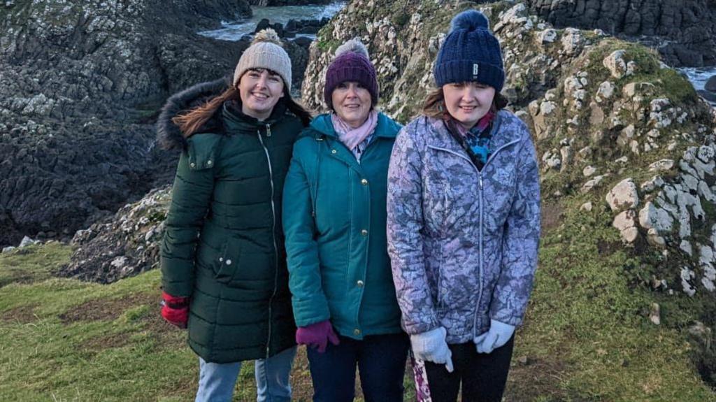 Two women and a teenager pose on top of rocks by the sea. They are all smiling and wearing bobble hats, gloves and coats. The sea is calm behind them.