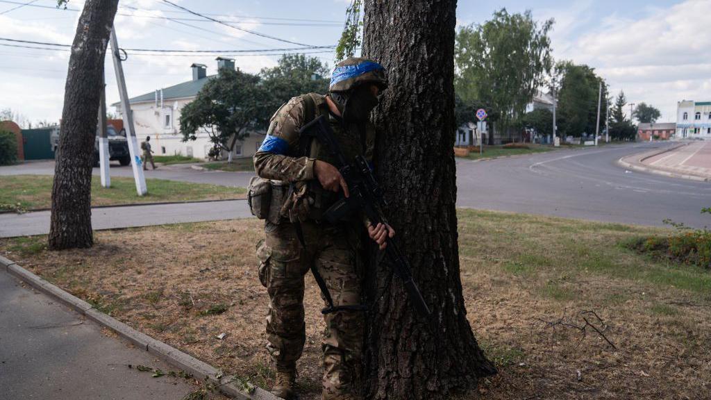 A Ukrainian soldier fighting in Russia's Kursk border region. He is carrying an assault rifle and is hiding behind a tree, while wearing a helmet and military fatigues. 