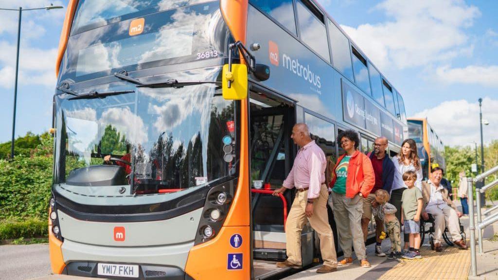 Picture of bright orange bus with the word 'Metrobus' on the side. There are people queuing to get on it.