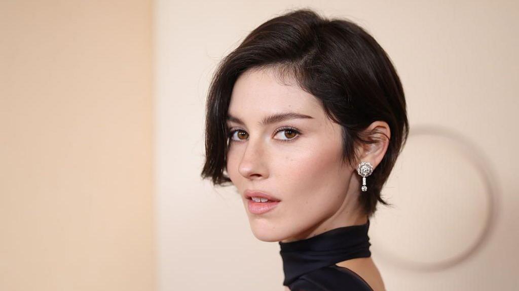 Gracie Abrams poses on the red carpet of the 2026 Oscars, in a black dress with a choker, and sparkling diamond earrings.