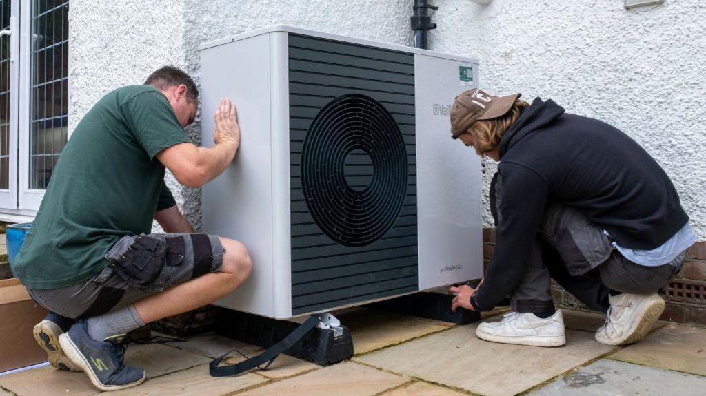 Two workers in casual clothing installing a heat pump - a large white box dominated by a black fan unit - on the outside a house with white harling walls