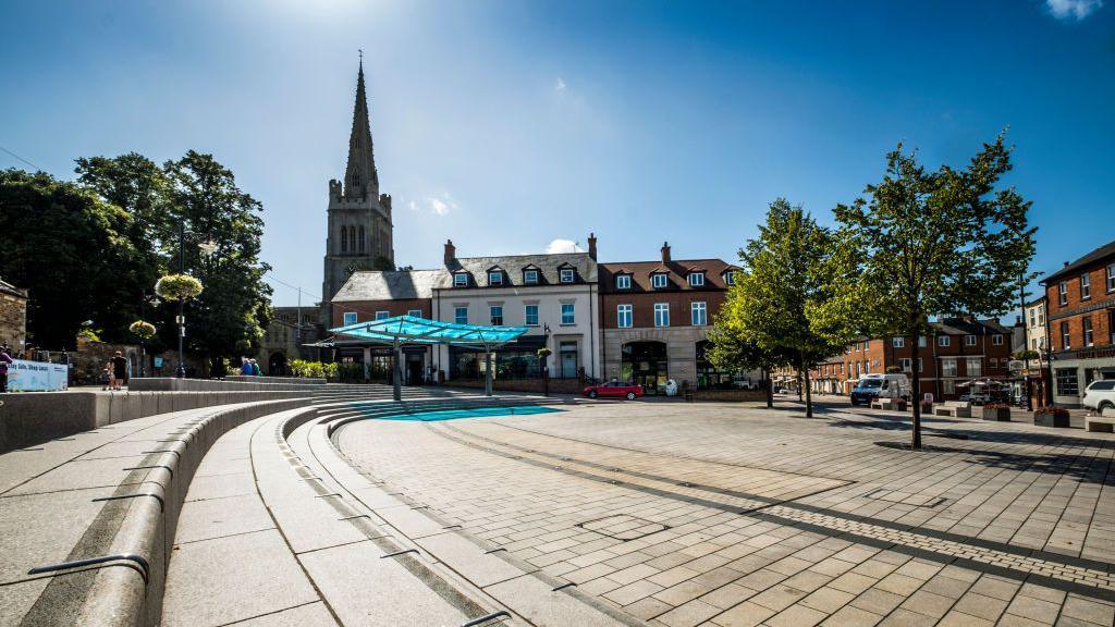 An empty concrete town centre square with a church spire and other houses  and buildings in the background. 