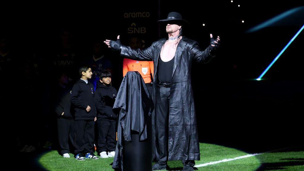 The Undertaker, in his trademark all-black outfit, makes his signature pose as two young children look on in awe