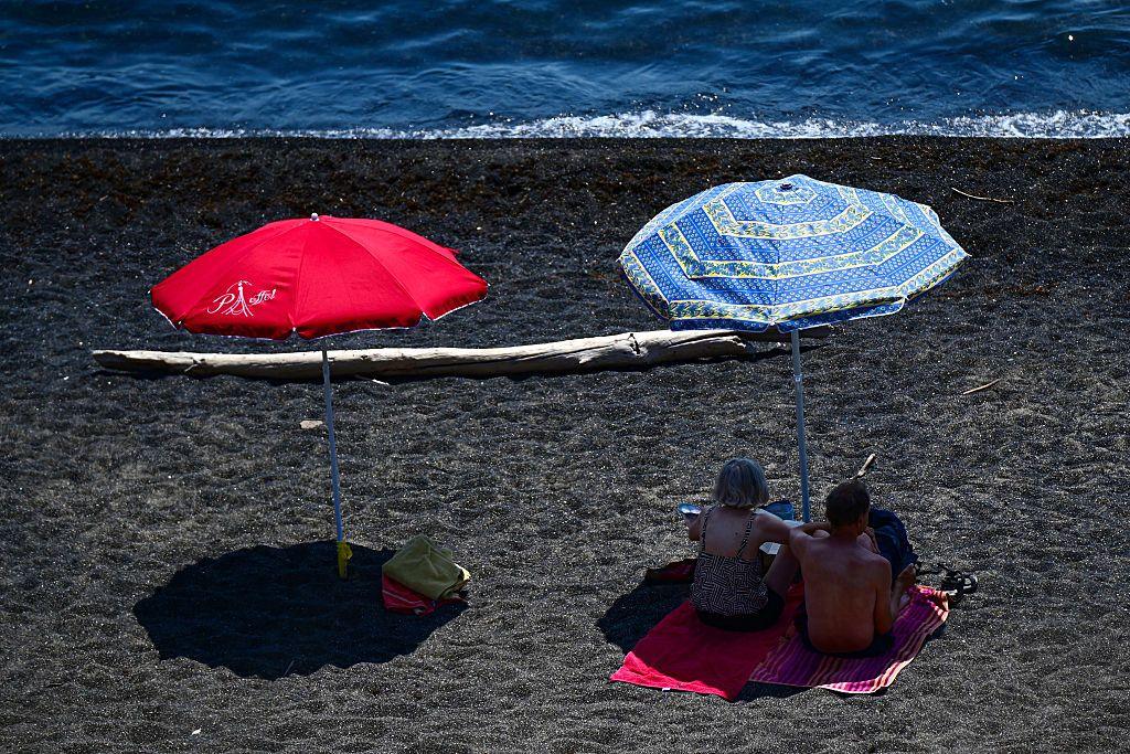 A red and blue parasol on a beach next to the sea, which is at the top of the picture. Two beachgoers are resting in the shade under the blue parasol on the right.
