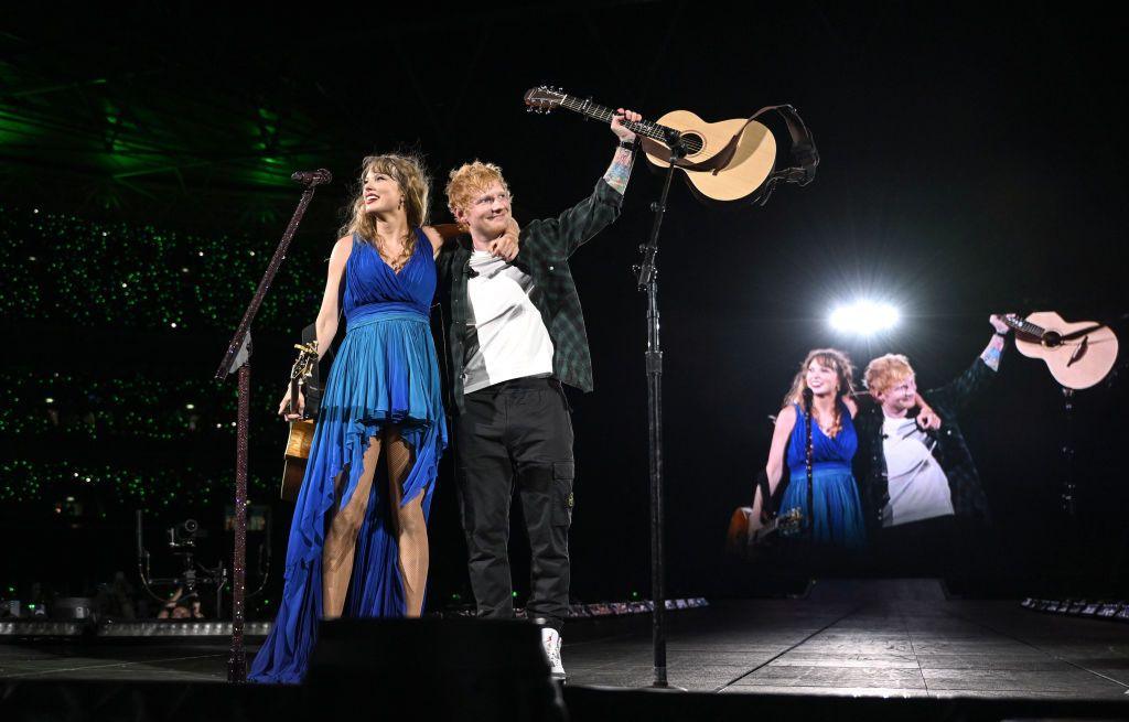 Taylor Swift and Ed Sheeran together on stage on the first night of concerts at London's Wembley stadium as part of her Eras tour. They are facing the crowd with their arms around one another. With his other arm, Sheeran is holding his guitar in the air.