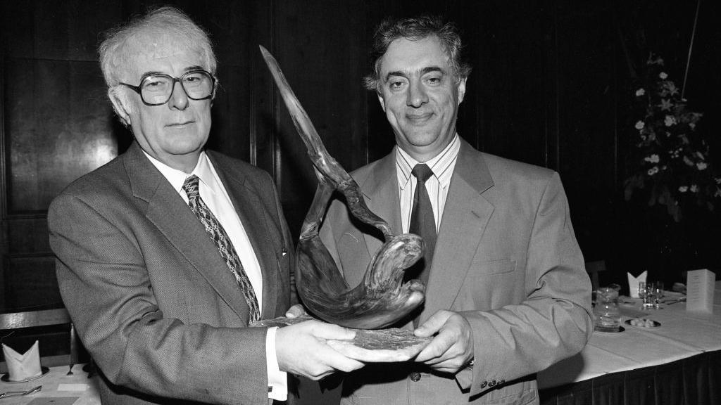 A black and white photo of a white haired man with black rimmed glasses, wearing a suit, being presented with a sculpture by a man with dark hair in a suit.