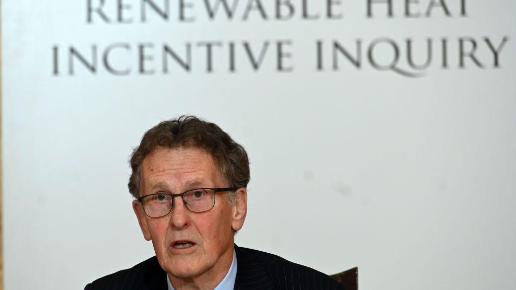 A dark haired man with dark rimmed glasses, sits with his mouth opening mid sentence. He wears a dark suit and blue shirt. The words Renewable heat incentive inquiry hang above him