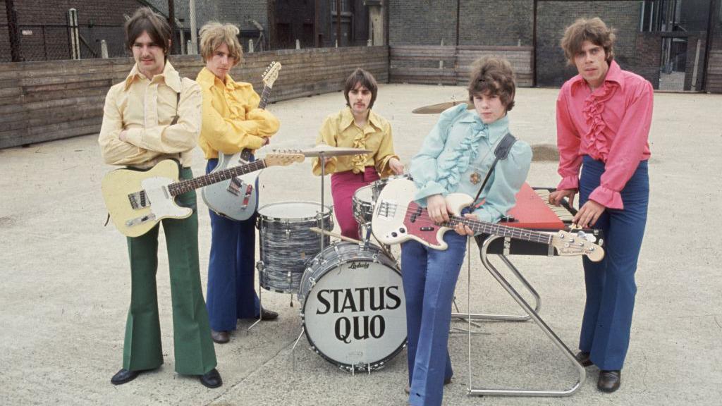 Status Quo, pictured in the 1960s looking very young, wearing flared jeans and brightly coloured shirts. They band have all their instruments and are posing outside with them