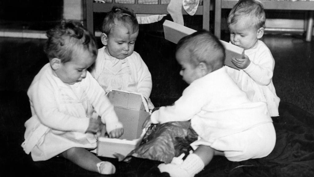 A black and white photograph shows four babies all dressed in white opening present while sitting on the floor