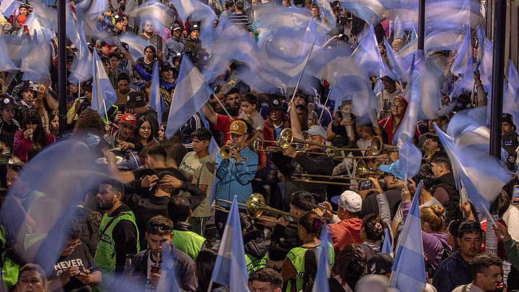 People play brass instruments in the middle of a crowd while others wave Argentinian flags