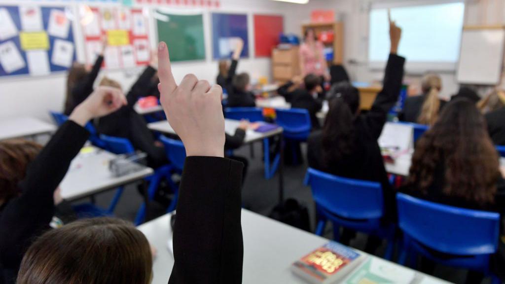 A blurry photo of a classroom of students with their backs to the camera, some of them with their hands up. There's a interactive whiteboard and a teacher in soft focus in front of them.