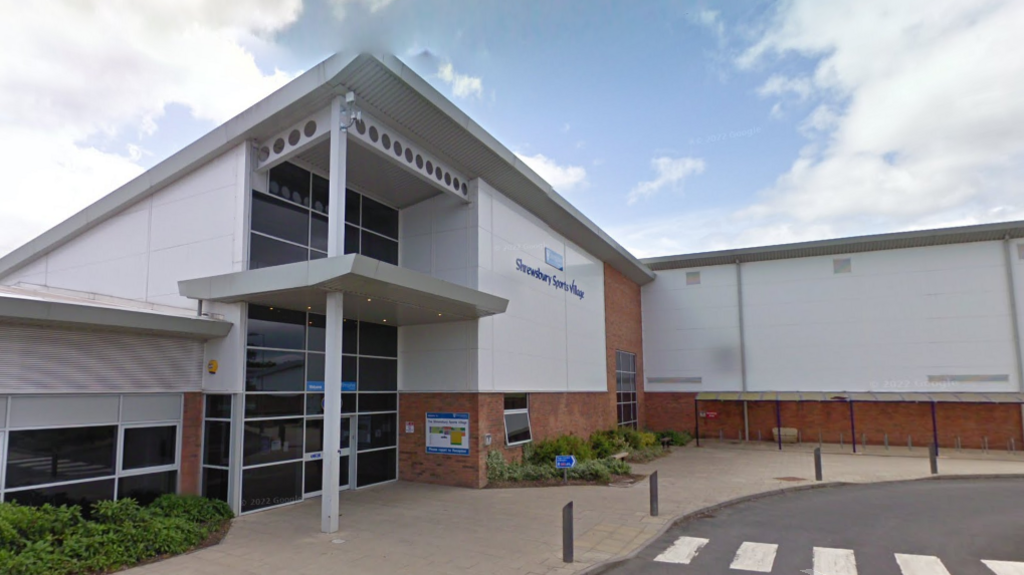 The front entrance of Shrewsbury Sports Village. It is a large white building with a pedestrian crossing in front of it.