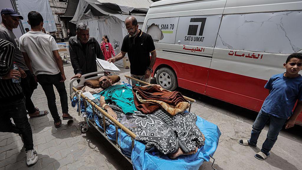 An injured man pictured outdoors on a bed on wheels, with other men in the background