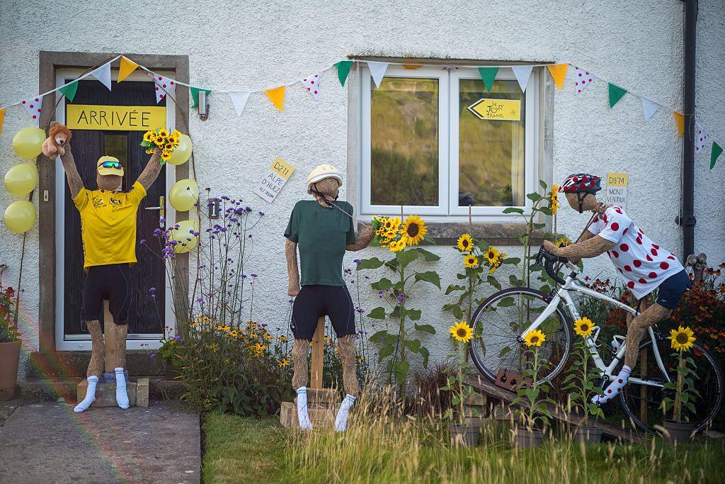 Three scarecrows are seen in front of a house. The house is decorated with bunting and a sign on the door which says "Arrivée" which means Arrived. One scarecrow is on a bike, while the others are seen cheering.