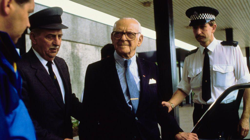 Armand Hammer, in blue shirt and and tie and dark suit, is being helped into a car by a police officer and a driver. They are all standing outside what appears to be a hospital.