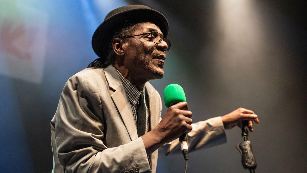 Neville Staple is wearing a black bowler hat and black glasses. He is wearing a beige blazer and is singing into a microphone. He is holding a black microphone stand in his other hand.