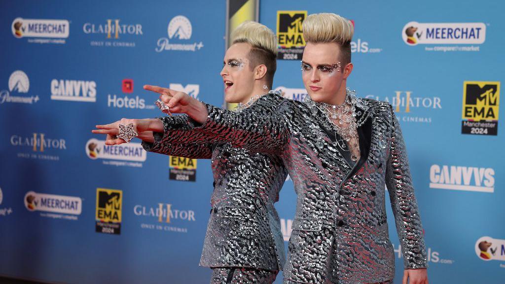 Jedward point at the camera in metallic suits in front of a blue background on the MTV Awards red carpet