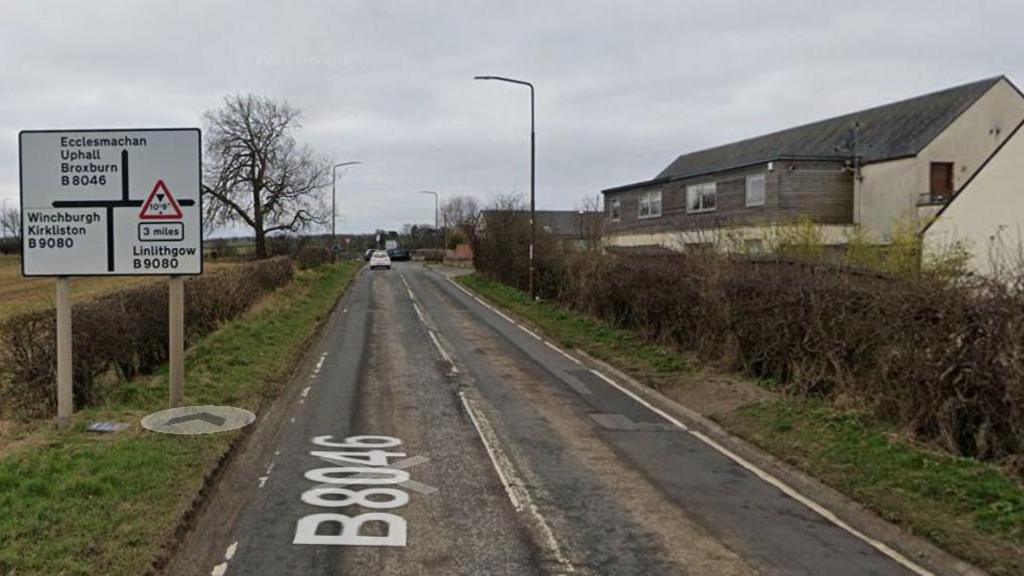 A road (the B8046). On the right hand side of the road is a row of houses, on the left is a field. A road sign points to Ecclesmachan, Linlithgow and Winchburgh. A white car is seen in the distance.