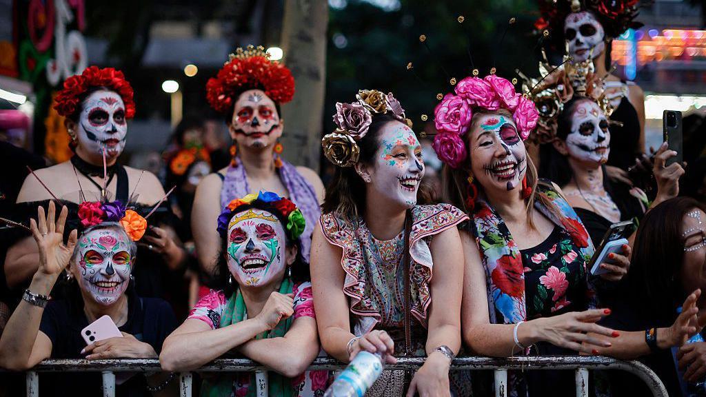 A group of women with their faces painted like skulls stand at a barrier, smiling and waving.