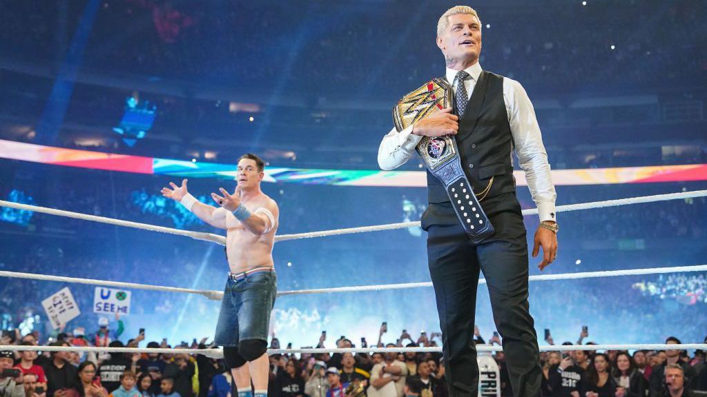 Cody Rhodes, a man in a suit and tie holding a champion wrestling belt stands in a wrestling ring while in the corner of the ring a man in long jean shorts and no top raises his hands.  