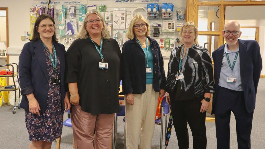 Five people are standing next to each other smiling at the camera. They are standing in front of a section in a library with medical equipment on the wall behind them.