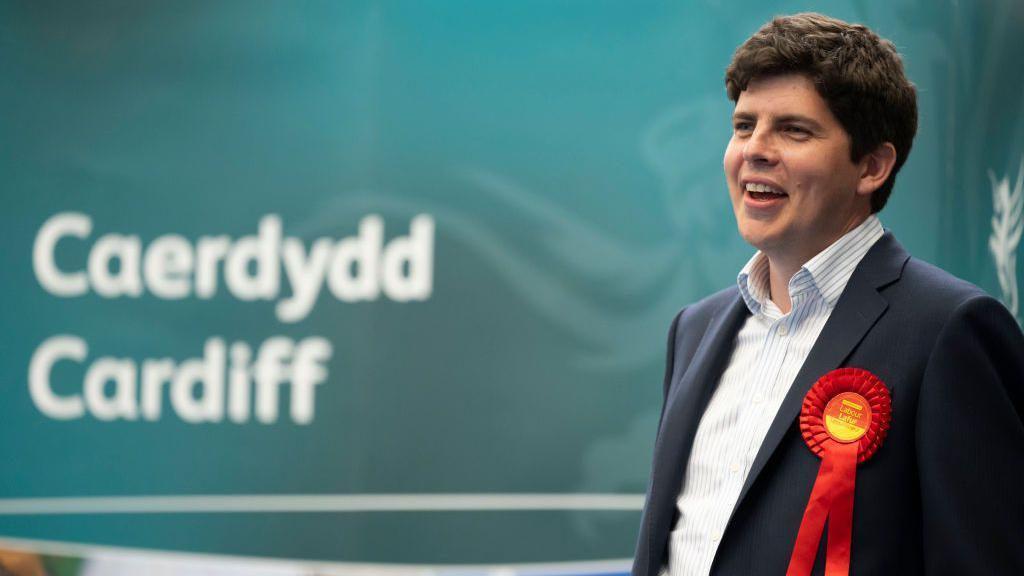 Huw Thomas, a dark haired man in a navy suit wearing a red rosette, stands in front of a large sign that says Caerdydd Cardiff