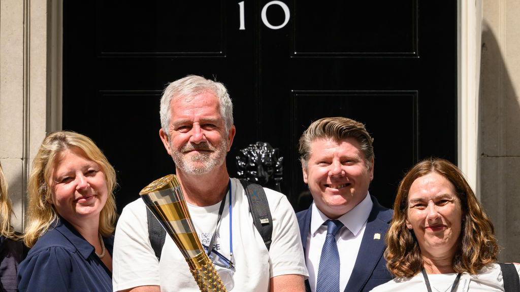 Two men and two women stood outside 10 Downing Street's black door. One man is wearing a white t-shirt and carrying a large gold and sliver baton.