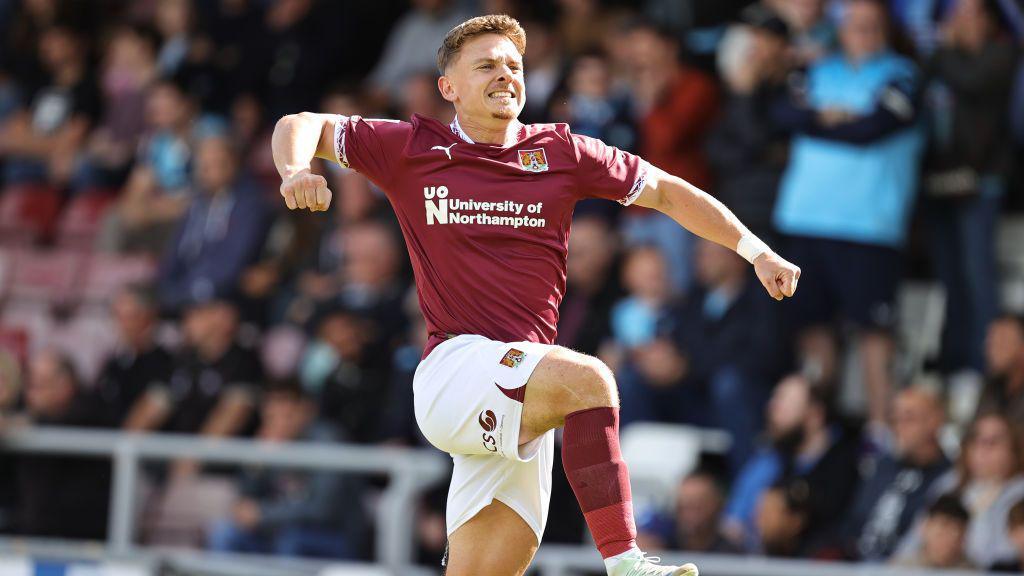 Sam Hoskins in a Claret Northampton Town kit jumping in the air to celebrate a goal in front of seated fans. He has short brown hair and is gritting his teeth.