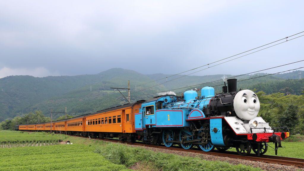 Thomas the tank engine replica pulls real yellow carriages along a track. Either side are green fields and hill in the background.
