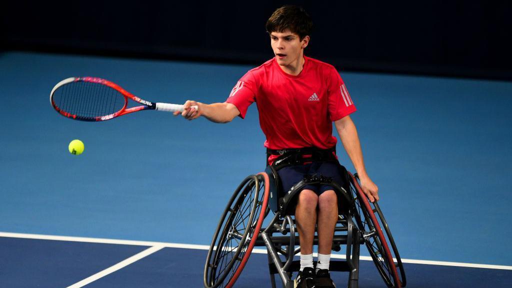 Greg Slade in a red shirt and wheel chair hitting a green tennis ball with a red tennis racket