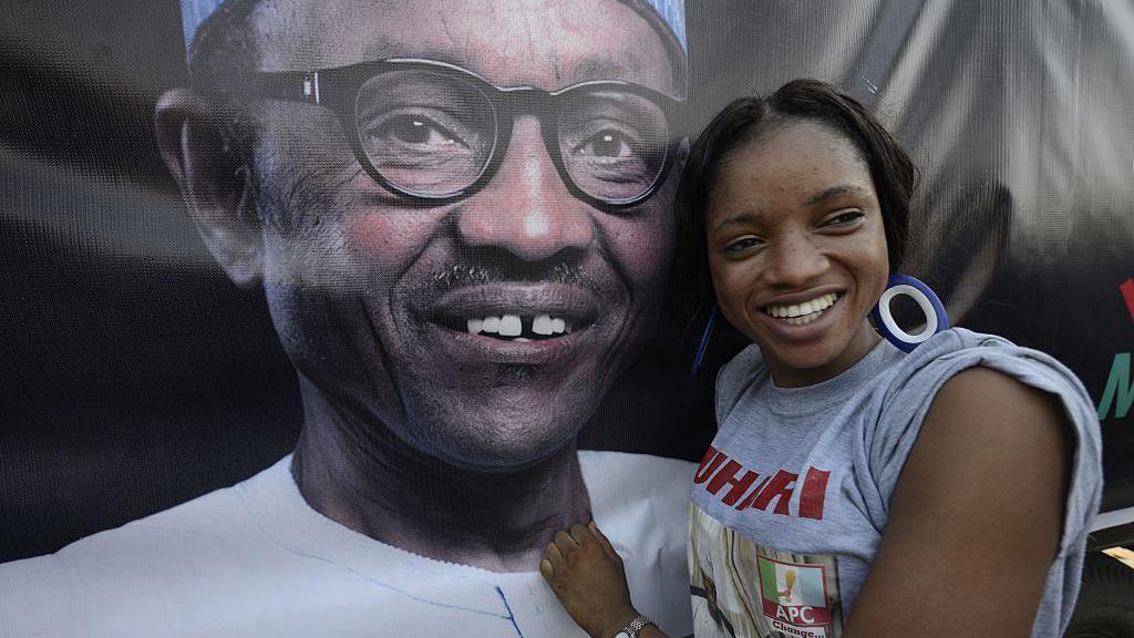 A woman poses next to a poster of a smiling Buhari. She wears a grey top written "APC" and "Buhari".