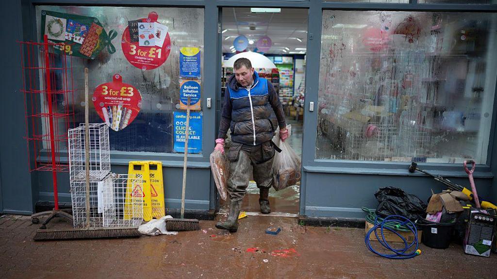 Dyn yn cerdded allan o'i siop gan gario bag llawn stoc.