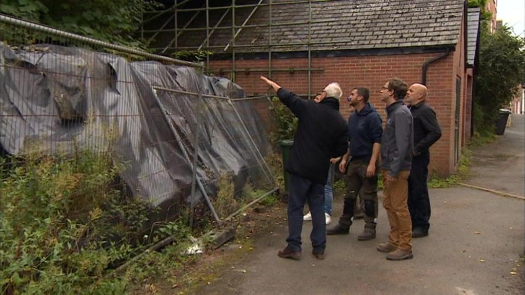 Six men standing, looking at a wall. The wall has been covered by black tarpaulin, and there are grey metal barriers in front of it.