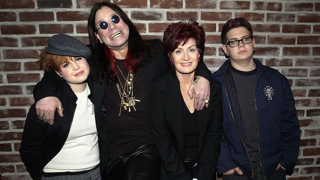 The Osbourne family pictured in front of a brick wall. Ozzy has his arms around Kelly on his right, and Sharon, left, and all three are smiling. Jack on the far left has his hands in his pockets and is not smiling. Ozzy is wearing all-black, black round sunglasses and heavy gold necklaces. His long hair is died black and red. Kelly's short hair is dyed red and she has a grey flat cap. Sharon is also all in black. Jack is wearing glasses, a navy jacket and grey jumper.