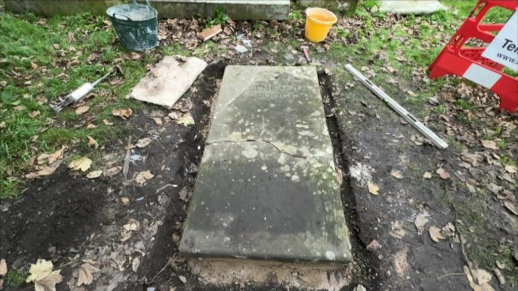 A repaired gravestone with cracks in it is lying on a concrete base on grass. The gravestone says "Ebenezer scrooge". There are buckets and equipment lying next to the stone