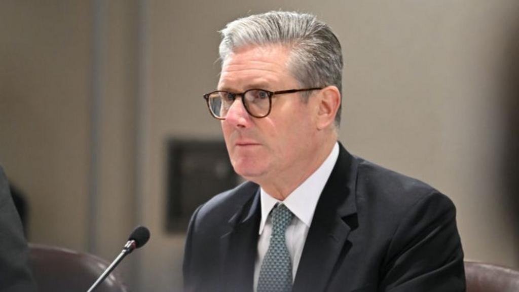 Keir Starmer sits down during a meeting to take questions. He's in a black suit, white shirt and patterned blue tie and is wearing glasses. The background is blurred