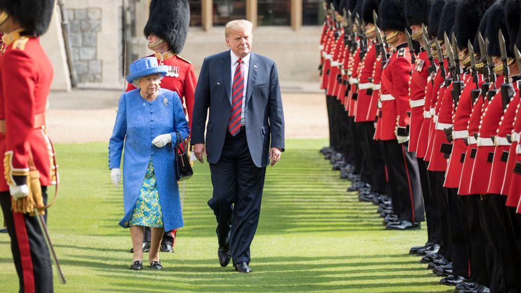Queen Elizabeth II and Donald Trump walk side by side on a neatly trimmed lawn, flanked by two rows of British Royal Guards in traditional red uniforms and tall black bearskin hats. The guards stand at attention with ceremonial rifles.