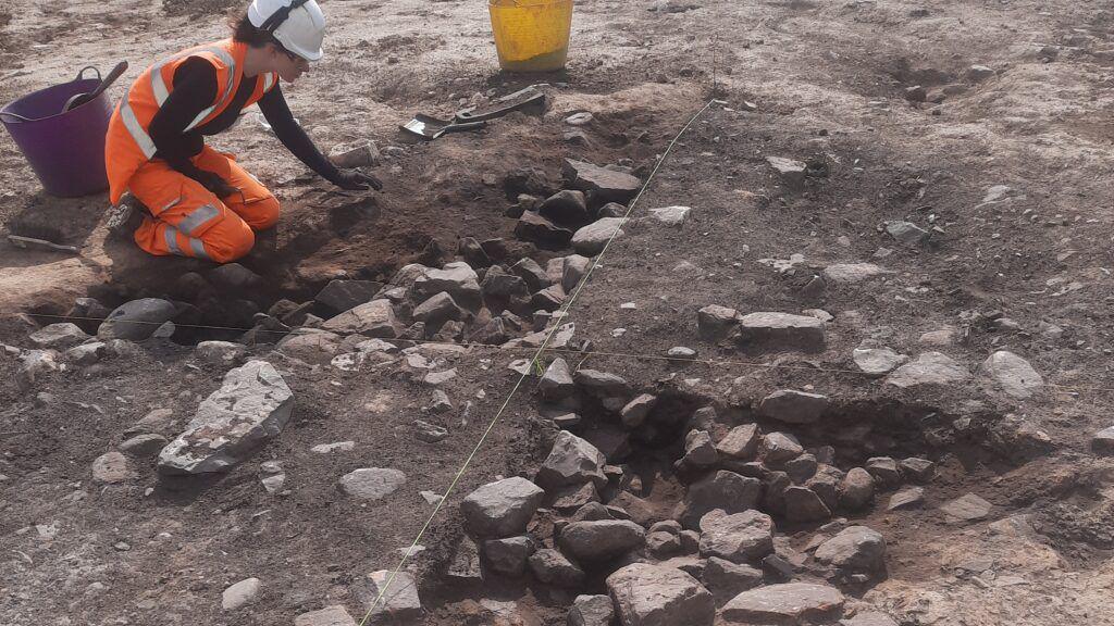 A young woman digs around rocks and mud in a pit. She is wearing orange high-vis jacket and trousers. There are buckets around her
