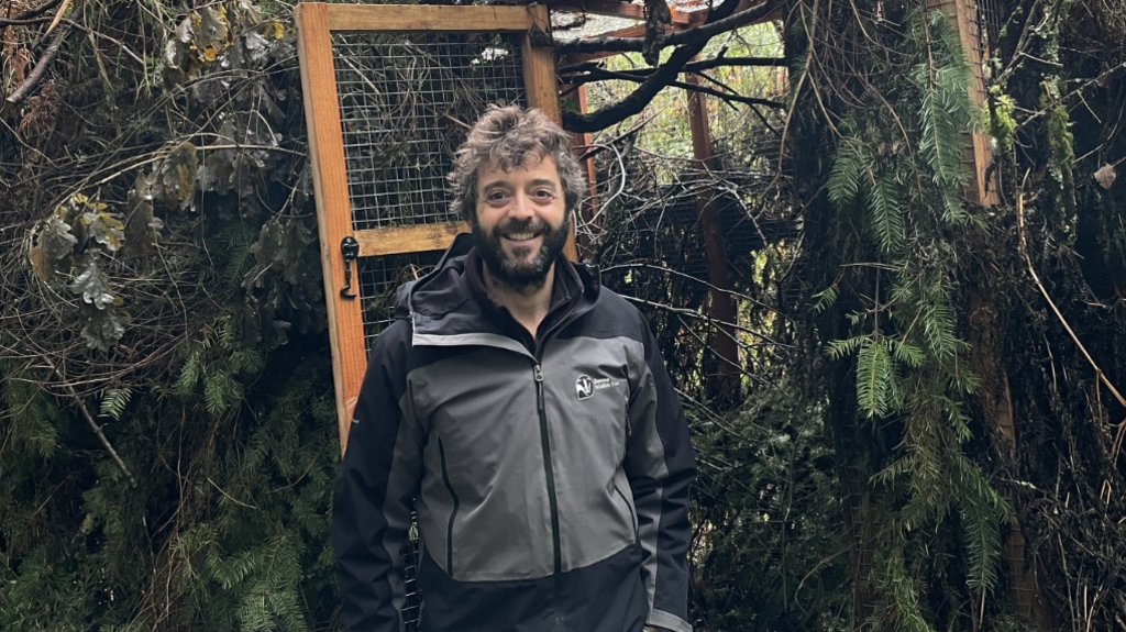 Ed Parr-Ferris stands in front of a large enclosure in woodland. He is wearing a grey and black Wildlife Trust jacket and is smiling at the camera. He has dark brown hair and a beard. 