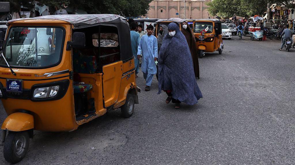 A woman covered with a long robe from head to toe, with her face covered by a medical mask, walks down the street near a tuktuk. Men can be seen in the background