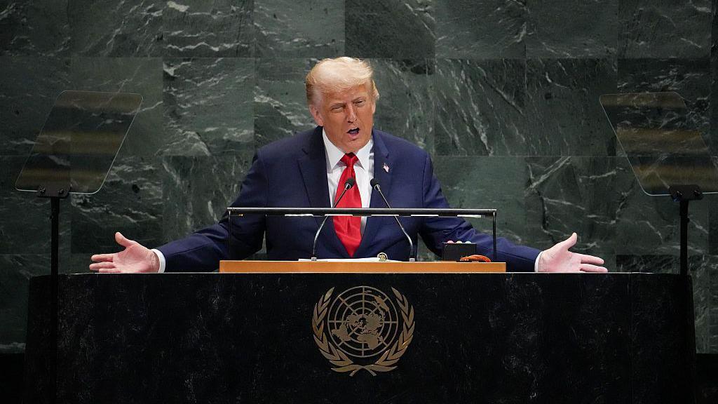 President Trump, in a navy blue suit, white shirt and red tie, stands behind a UN lectern giving a speech. He has both arms outstretched.