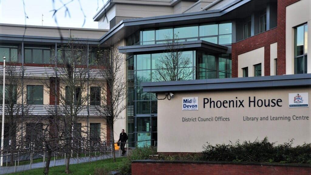 The photograph shows the outside of Phoenix House, Tiverton. In the foreground is a sign that reads "Phoenix House, District Council offices". The building behind the sign has a glass entrance and the building appears to be three to four storeys high.