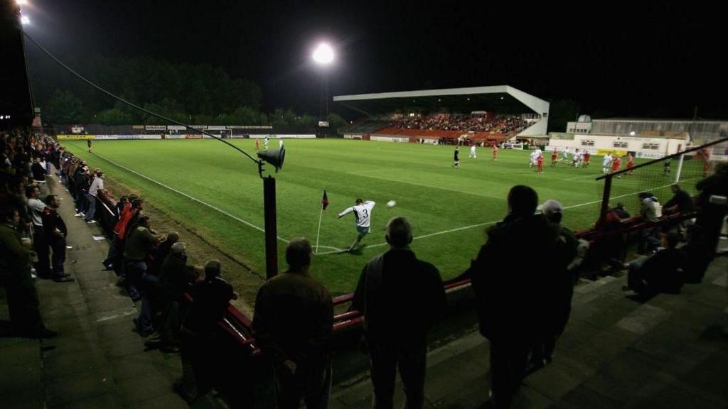 Kettering Town's Rockingham Road stadium during a evening match. A crowd is watching on as a player in a white shirt takes a corner kick.