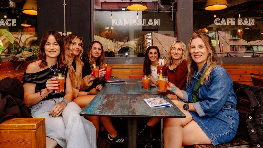 A group of six young women sit around a booth in a bar in Bristol, all of them holding red cocktails in glasses and smiling at the camera.