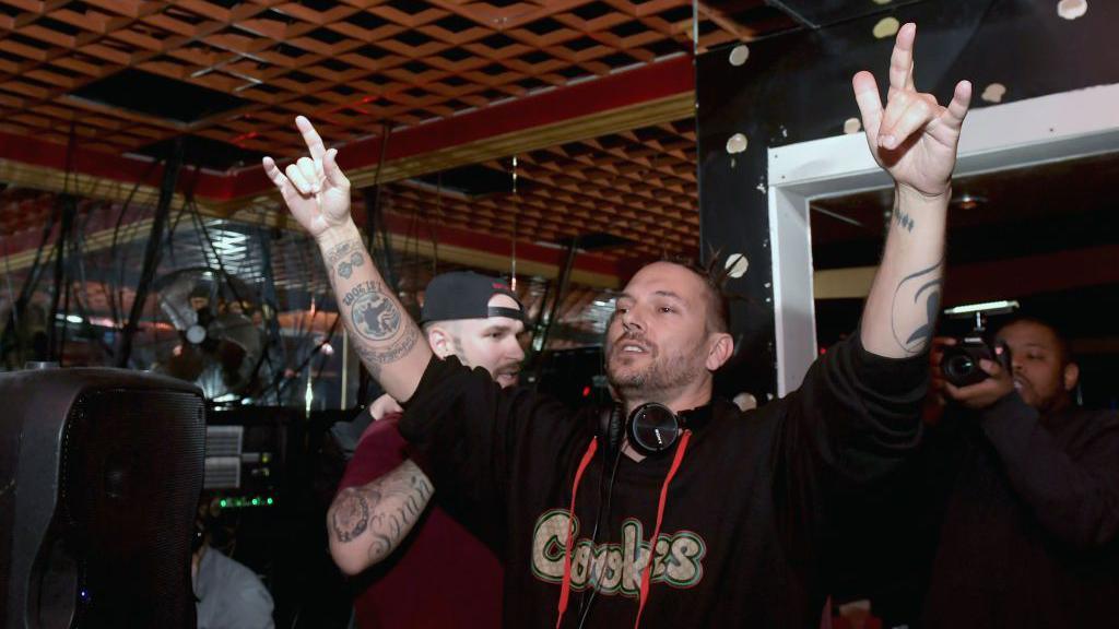 Federline, wearing headphones at the DJ booth, raises his hands in the air