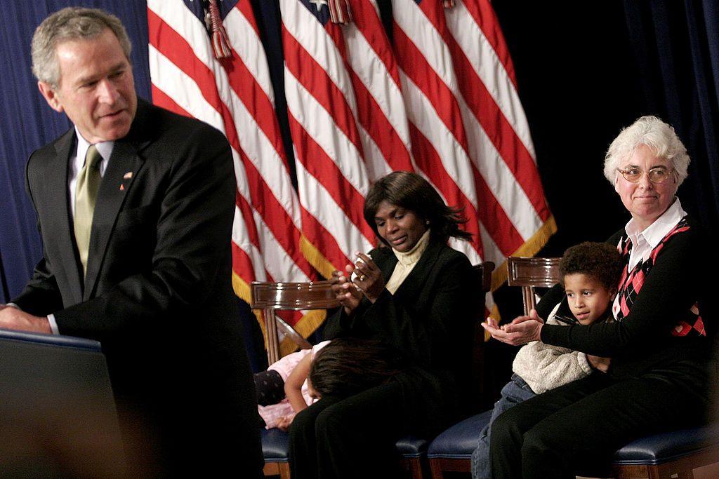 Standing at a podium, George W. Bush, in a suit, turns his head as South Africa's Thandazile Darby and Dr Helga Holst, both seated with children, applaud on 1 December 2005 as World Aids Day is commemorated in the Eisenhower Executive Office Building in Washington, DC.