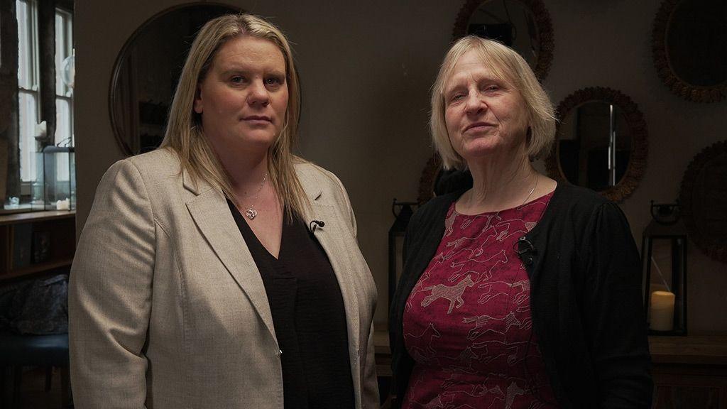 Micala and Sue stand side by side looking defiant. Micala has long blond hair and is wearing a black shirt and light brown jacket. Sue has short blond hair and is wearing a red top with brown horses on it and a black jacket. Behind them is a wall with several mirrors on it 