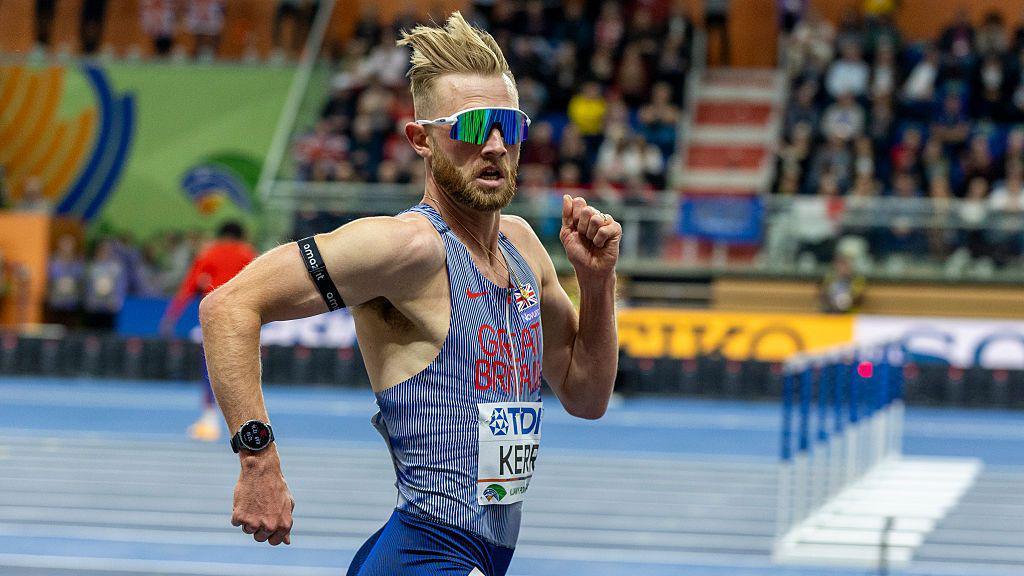 Josh Kerr's blonde hair is pushed back as he runs on the track, looking at the camera. 