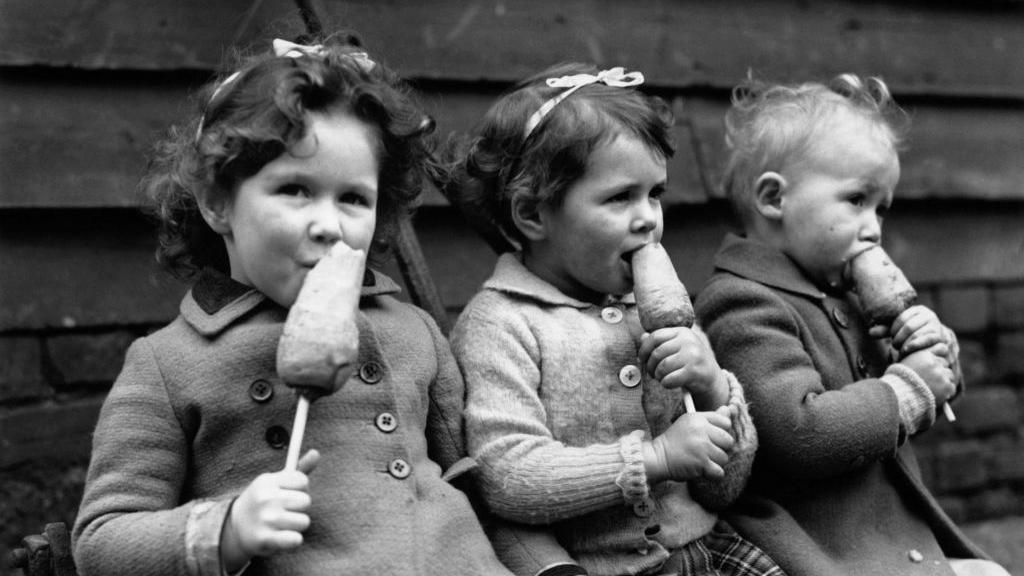 Black-and-white photo of three children sitting on a bench eating some kind of vegetable, possibly carrots, on a stick.